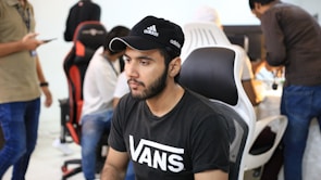 A group of young people are working in a modern office setting with computers. The focus is on a man wearing a black Adidas cap and a black Vans T-shirt, seated on a black and white chair. Others in the background also work on computers, with some standing and one holding a smartphone.