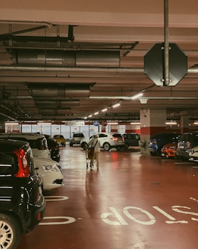 An indoor parking garage with multiple cars parked in rows. The floor has the word 'STOP' painted on it. A person is pushing a shopping cart toward the back exit, where there is a sign marked 'USCITA'. The lighting is warm, with industrial ceiling fixtures.