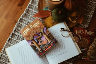 A cozy arrangement featuring an open book with a Harry Potter cover placed on it, surrounded by autumnal decorations like pumpkins, leaves, and a candle. A pair of glasses rests casually on top of the book.