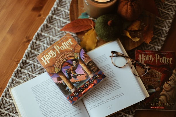 A cozy arrangement featuring an open book with a Harry Potter cover placed on it, surrounded by autumnal decorations like pumpkins, leaves, and a candle. A pair of glasses rests casually on top of the book.