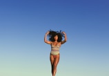 A person in a colorful, striped swimsuit poses confidently against a clear blue sky. The individual stands with arms raised, embracing their natural curly hair, conveying a sense of freedom and self-expression.