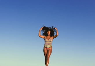 A person in a colorful, striped swimsuit poses confidently against a clear blue sky. The individual stands with arms raised, embracing their natural curly hair, conveying a sense of freedom and self-expression.