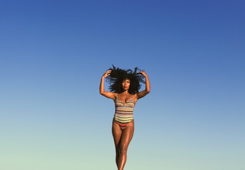 A person in a colorful, striped swimsuit poses confidently against a clear blue sky. The individual stands with arms raised, embracing their natural curly hair, conveying a sense of freedom and self-expression.