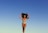 A person in a colorful, striped swimsuit poses confidently against a clear blue sky. The individual stands with arms raised, embracing their natural curly hair, conveying a sense of freedom and self-expression.