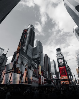Skyscrapers rise against a cloudy sky, adorned with bright, large advertisements and digital billboards. The urban environment is dynamic, with various colorful ads promoting different brands and shows.