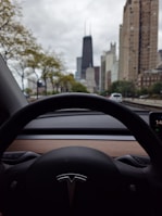 View from inside a car, specifically a Tesla, with the steering wheel prominently in the foreground. The windshield reveals a city landscape with tall skyscrapers and trees lining the street. The weather appears cloudy, and there are a few cars on the road.