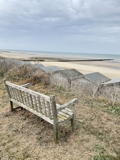 A weathered wooden bench is situated on a grassy hillside overlooking a beach. In the background, there are several beach huts and a vast expanse of sand leading to the sea. The sky is overcast, with a mix of gray and white clouds.