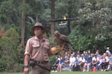 A person wearing a brown uniform and hat is holding a bird of prey on their gloved hand in an outdoor setting surrounded by lush greenery. Another bird is flying in the background. A group of people are sitting and watching the scene unfold.