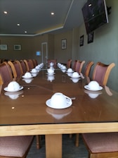 A long, polished wooden conference table is set with multiple white teacups and saucers, positioned in front of each chair along the table's perimeter. The room's walls are adorned with framed photographs and a flat-screen television mounted on the right side. The lighting is soft, with recessed ceiling lights. The seating consists of cushioned chairs with wooden frames, all facing inward towards the table.