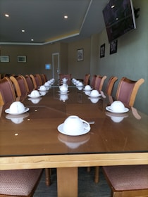 A long, polished wooden conference table is set with multiple white teacups and saucers, positioned in front of each chair along the table's perimeter. The room's walls are adorned with framed photographs and a flat-screen television mounted on the right side. The lighting is soft, with recessed ceiling lights. The seating consists of cushioned chairs with wooden frames, all facing inward towards the table.