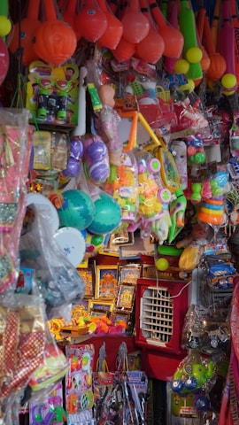 A densely packed display of assorted colorful toys hanging and stacked in a store. The items include various types of balls, rackets, action figures, dolls, and other children's playthings in vibrant packaging.