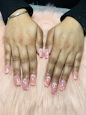 Hands with freshly manicured nails featuring a glossy peach base color decorated with intricate floral patterns. The background consists of a soft pink, fluffy surface.