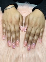 Hands with freshly manicured nails featuring a glossy peach base color decorated with intricate floral patterns. The background consists of a soft pink, fluffy surface.