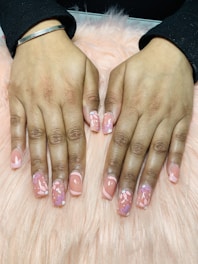 Hands with freshly manicured nails featuring a glossy peach base color decorated with intricate floral patterns. The background consists of a soft pink, fluffy surface.