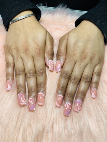 Hands with freshly manicured nails featuring a glossy peach base color decorated with intricate floral patterns. The background consists of a soft pink, fluffy surface.