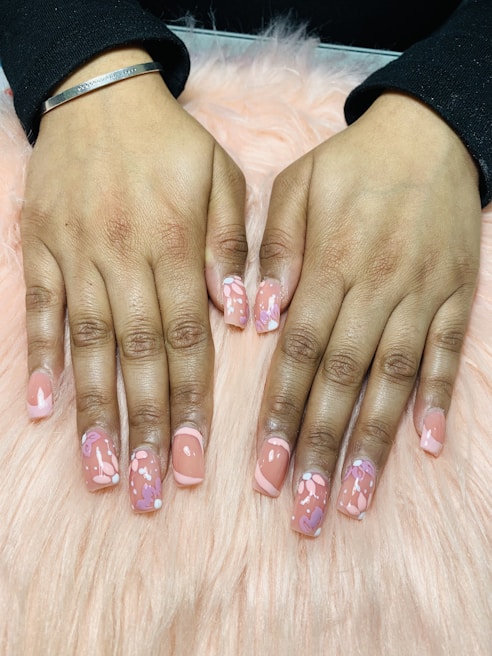 Hands with freshly manicured nails featuring a glossy peach base color decorated with intricate floral patterns. The background consists of a soft pink, fluffy surface.