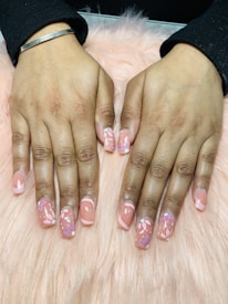 Hands with freshly manicured nails featuring a glossy peach base color decorated with intricate floral patterns. The background consists of a soft pink, fluffy surface.