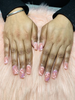 Hands with freshly manicured nails featuring a glossy peach base color decorated with intricate floral patterns. The background consists of a soft pink, fluffy surface.