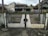 A metal gate with vertical black bars and white panels, showing signs of rust and wear, stands in front of a paved street. Behind it, there are several houses with peaked roofs and weathered walls, surrounded by small patches of greenery.
