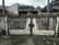 A metal gate with vertical black bars and white panels, showing signs of rust and wear, stands in front of a paved street. Behind it, there are several houses with peaked roofs and weathered walls, surrounded by small patches of greenery.