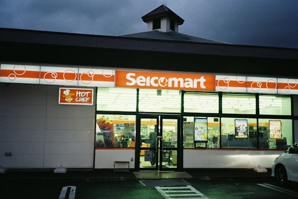 A convenience store with a bright orange and white sign displaying the name 'Seicomart'. The store's exterior features large glass windows through which various products and advertisements are visible. The entrance is well-lit, and a portion of a vehicle is parked in front.