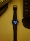 A smartwatch with a black strap lies on a surface, displaying a digital watch face with various dials and information. To its right, part of a computer keyboard is visible, while the upper left corner shows part of a cylindrical object.