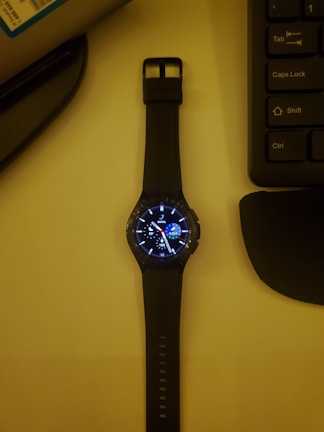 A smartwatch with a black strap lies on a surface, displaying a digital watch face with various dials and information. To its right, part of a computer keyboard is visible, while the upper left corner shows part of a cylindrical object.
