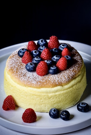A fluffy, golden-brown cake topped with a dusting of powdered sugar, garnished with fresh raspberries and blueberries. The cake is placed on a white plate against a dark background, highlighting its texture and vibrant colors of the fruits.