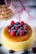 A fluffy, golden-brown cheesecake is topped with vibrant blueberries and raspberries. The cake sits elegantly on a white plate, with additional berries scattered around. In the background, a decorative woven basket and soft focus pink flowers add a touch of warmth and elegance.