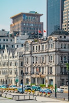 Urban scene featuring historic and modern buildings with intricate architecture. The area is populated with cars, suggesting a busy city environment. Flags are visible atop one of the buildings, adding a sense of identity.