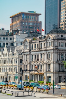 Urban scene featuring historic and modern buildings with intricate architecture. The area is populated with cars, suggesting a busy city environment. Flags are visible atop one of the buildings, adding a sense of identity.