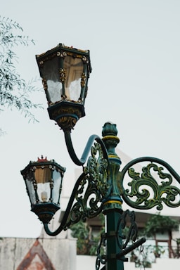An ornate, antique-style street lamp featuring intricate metalwork and three lanterns with decorative glass panels. A leafy tree branch is visible on the left side and parts of buildings are in the background.