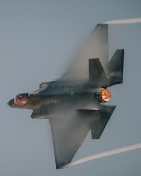 A military jet flies at high speed, producing vapor trails and emitting bright flames from its engine. The jet exhibits a sleek and aerodynamic design.