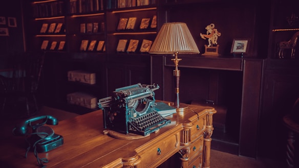 An elegant vintage study room with a wooden desk at the center featuring an antique typewriter and a green rotary phone. The background shows bookshelves filled with books and various ornaments, including a decorative statue and an old lamp with a pleated shade. The ambiance is warm and inviting, with a cozy and nostalgic feel.