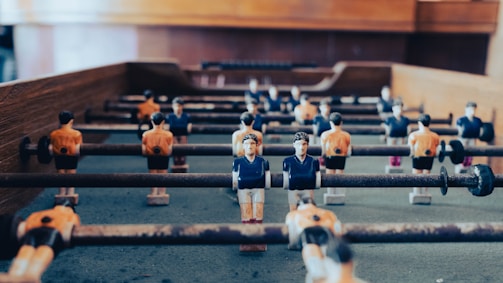 A close-up view of a foosball table with miniature soccer player figures attached to rotating rods. The figures are dressed in blue and orange jerseys, positioned in offensive and defensive formations. The table surface is worn, with the surrounding wooden frame visible.