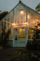 A warmly lit glass greenhouse during twilight, surrounded by foliage. Soft lighting creates an inviting glow from within, highlighting the glass panels and entrance with two wall-mounted lamps.