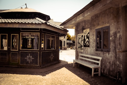 A rustic outdoor scene featuring two adjacent buildings with vintage architecture. The building on the left has signs indicating it is a tattoo studio, with decorative star motifs on the walls. On the right, a weathered bench sits below posters on a rough-textured wall. The area is illuminated by bright sunlight, casting shadows on the cobblestone path.