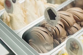 A gelato display featuring two flavors in metallic trays. One is labeled 'Double Dark Chocolate' with dark brown swirls, while the other is a light cream color, likely vanilla or a similar flavor. The presentation is enticing and neatly arranged.