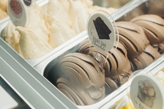 A gelato display featuring two flavors in metallic trays. One is labeled 'Double Dark Chocolate' with dark brown swirls, while the other is a light cream color, likely vanilla or a similar flavor. The presentation is enticing and neatly arranged.