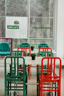 An indoor setting featuring a minimalist dining area with tables and chairs in vibrant colors. The chairs are predominantly green and red, and each table has a small green plant as decoration. The area is well-lit by natural light coming through large windows with frosted glass and some signage visible.