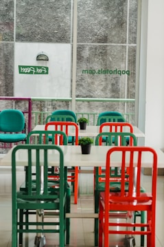 An indoor setting featuring a minimalist dining area with tables and chairs in vibrant colors. The chairs are predominantly green and red, and each table has a small green plant as decoration. The area is well-lit by natural light coming through large windows with frosted glass and some signage visible.