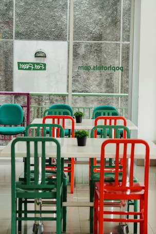 An indoor setting featuring a minimalist dining area with tables and chairs in vibrant colors. The chairs are predominantly green and red, and each table has a small green plant as decoration. The area is well-lit by natural light coming through large windows with frosted glass and some signage visible.