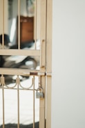A metal gate with vertical bars and decorative elements is secured with a padlock. The gate is a light beige color and is attached to a white wall. In the background, there is a blurred view of what appears to be a shadowed interior space with some indistinct objects.