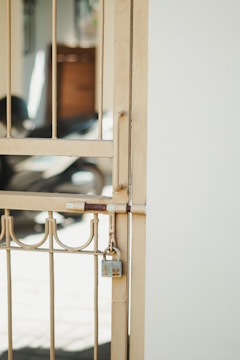 A metal gate with vertical bars and decorative elements is secured with a padlock. The gate is a light beige color and is attached to a white wall. In the background, there is a blurred view of what appears to be a shadowed interior space with some indistinct objects.