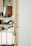 A metal gate with vertical bars and decorative elements is secured with a padlock. The gate is a light beige color and is attached to a white wall. In the background, there is a blurred view of what appears to be a shadowed interior space with some indistinct objects.