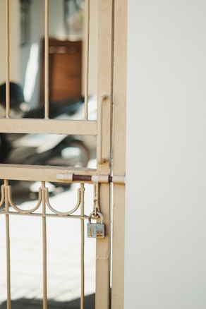 A metal gate with vertical bars and decorative elements is secured with a padlock. The gate is a light beige color and is attached to a white wall. In the background, there is a blurred view of what appears to be a shadowed interior space with some indistinct objects.