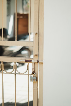A metal gate with vertical bars and decorative elements is secured with a padlock. The gate is a light beige color and is attached to a white wall. In the background, there is a blurred view of what appears to be a shadowed interior space with some indistinct objects.
