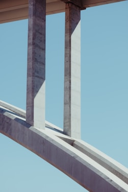 Concrete bridge structure featuring large vertical pillars and an arch against a clear blue sky. The geometry of the bridge creates interesting shadows and lines.