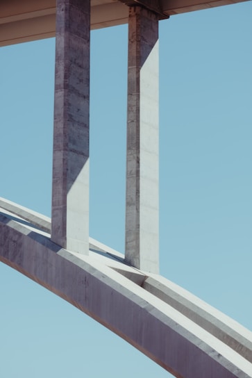 Concrete bridge structure featuring large vertical pillars and an arch against a clear blue sky. The geometry of the bridge creates interesting shadows and lines.