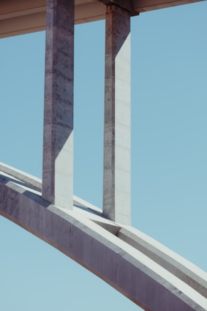 Concrete bridge structure featuring large vertical pillars and an arch against a clear blue sky. The geometry of the bridge creates interesting shadows and lines.